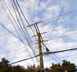 Poste de luz amarillo lleno de cables con un cielo azul y nubes de fondo