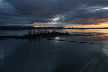 The riverbank and small island in the Columbia river connecting Portland, Oregon with Pacific Ocean overgrown by trees in winter season during sunset. In background are mountains under dramatic clouds