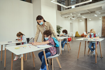 Modern teacher wearing protective face mask helping little boy during lesson. Kids studying in elementary school, sitting at the desk