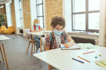 School boy wearing face mask during corona virus and flu outbreak writing in his notebook while sitting at the desk in a classroom. Kids studying in elementary school
