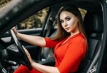 Obraz na płótnie Canvas A beautiful young girl in a red overalls sits behind the wheel of a black car on an empty road in the forest