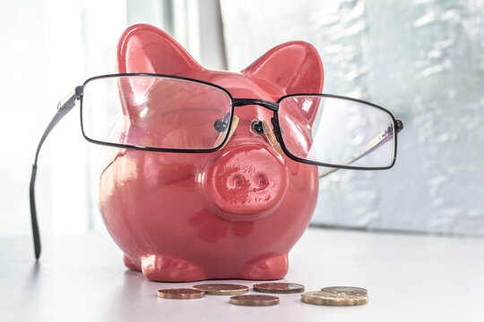 A piggy bank with glasses stands by the window, next to it are several euro coins. Slightly fun and sloppy scene with the concept of saving money, saving, spending, shopping