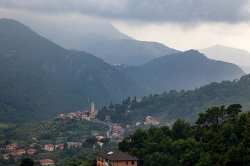 Fototapeta na wymiar Beautiful views of the Italian village in the mountains. Temples and rooftops among the greenery. Low clouds after rain.