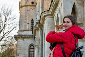 Fototapeta na wymiar A girl in a red jacket holds a ginger cat in her arms standing next to the wall of an old building