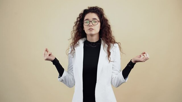 The young woman with calm and calm curly hair holds her hands sideways in the form of yoga, relaxing. Young hipster in gray jacket and white shirt, with glasses posing isolated on beige background in