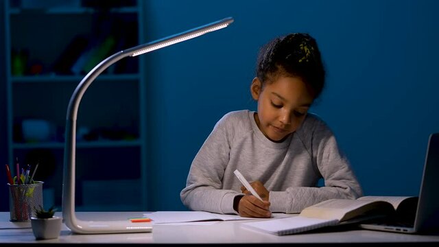 The girl rewrites the task from the textbook into a notebook. A schoolgirl sits at a table by the light of a night lamp. Homework, distance learning. Close up. Slow motion ready 59.97fps.