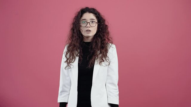 The young woman balanced with curly hair, holding her finger to her lips shows the sign of silence. Young hipster in white jacket and black shirt, with glasses posing isolated on pink background in