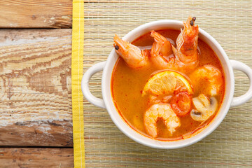 Tom yam soup with shrimps in a tureen on a mat on a wooden table.