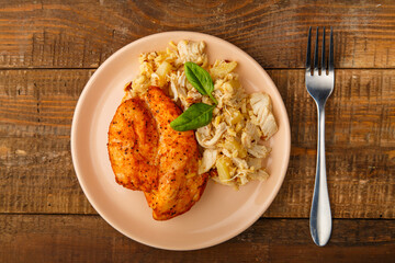 Baked chicken breast with salad on a plate next to a fork on a wooden table.