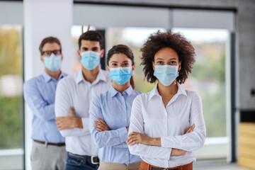 Mixed race businesswoman with face mask standing in corporate firm with arms crossed. Behind her are her colleagues with arms crossed and face masks, too. Although it's corona business can't stop.