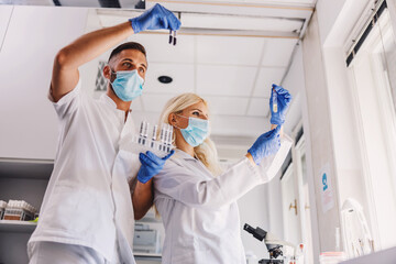 Two dedicated lab assistants with rubber gloves and face masks standing in laboratory and holding test tubes during corona virus outbreak.