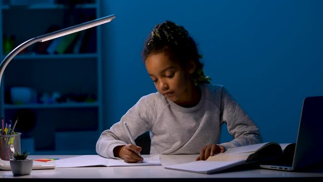 A girl rewrites homework, yawns widely, rests her head on her hands, falls asleep. The child sits at a table by the light of a night lamp. Close up. Slow motion ready 59.97fps.