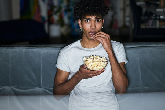 Frightened Young Guy With Eyes Wide Open Wearing Pajamas Eating Popcorn While Watching Horror Movie, Sitting On A Comfortable Couch At Home