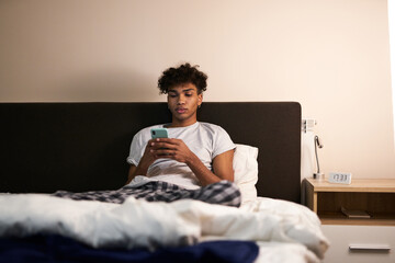 Young guy with piercing chatting with friends, using smartphone in his bed at night before going to...