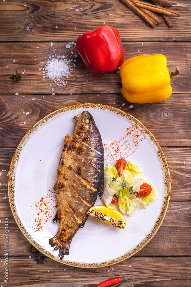 Wall mural Grilled fish on a plate with vegetables, bell pepper, coarse salt on a brown wooden background.