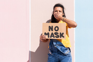 Portrait of hispanic woman holding a banner with the text No Mask while covering her mouth with her hand. She is standing next to a colorful wall. Space for text.