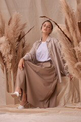 Young elegant female model sitting at beige textile background and posing between ears of rye. Brunette girl with makeup and pony tail hair in white shirt and beige skirt and jacket