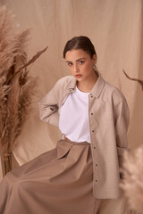 Young elegant female model sitting at beige textile background and posing between ears of rye. Brunette girl with makeup and pony tail hair in white shirt and beige skirt and jacket