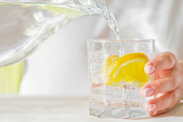 Female hands pouring water from the decanter into a glass beaker with lemon and ice. Health and...