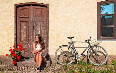 Fototapeta premium Quizzical girl sits outside her faded wooden door next to her bicycle in this retro hipster image