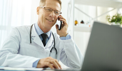 Cheerful doctor talking on cellphone and using laptop in clinic