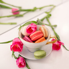 Sweet dessert, macaroons in golden mug and pink tulips on white wool background