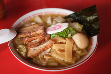 Asian noodle soup, ramen with roast pork, bamboo shoot, vegetables, seaweed, and boiled egg in a white bowl. Red table background, selective focus.