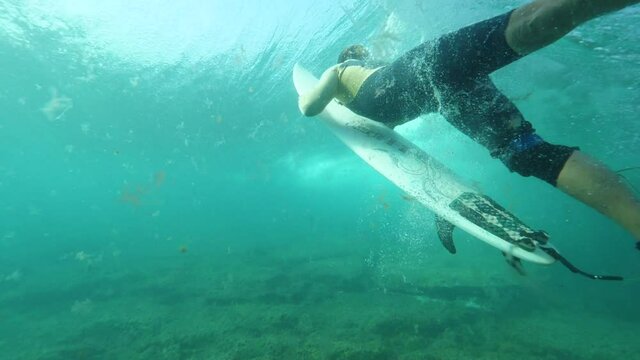 UNDERWATER, SLOW MOTION, CLOSE UP: Unknown surfer dude paddling on cool surfboard to catch a wave. Extreme surfer doing a duck dive in crystal clear ocean water. Surfer paddling out of a deep wave.