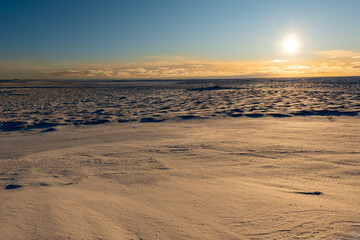 A bright orange sunset over snowy barren land. There are trees and shrubs in the distance with snow in the foreground. The sky is orange with thick clouds and an orange ball near the horizon. 