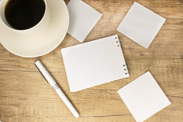 A notebook with a blank sheet of paper lies on a wooden table among white blank sheets and a white cup of coffee.