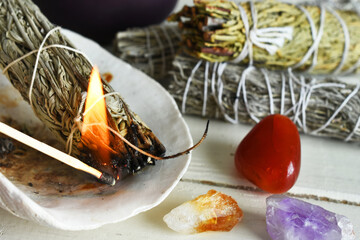 A close up image of a burning white sage smudge stick and healing crystals on a white background. 