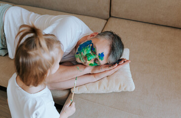 A little girl paints her father's face with a brush and watercolors while he sleeps on the couch