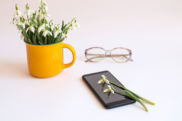 A bouquet of snowdrops in a yellow cup, a phone with two snowdrops, glasses for vision on a white background