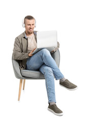 Young man with laptop listening to music while sitting in armchair on white background