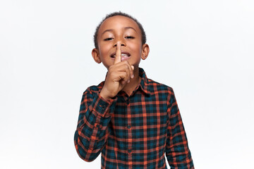 Isolated shot of adorable African boy holding fore finger on his lips, saying shh, asking for silence. Cute black child making gesture at mouth, whispering, shushing. Be quiet. Don’t speak so loud