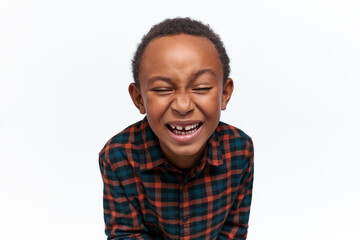 Funny adorable African little boy wearing stylish plaid shirt having cheerful look, closing eyes,...