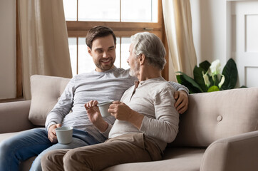 Happy millennial adult Caucasian man relax on sofa in living room with senior father talk drink...