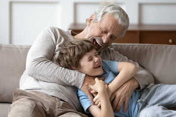 Overjoyed older Caucasian grandfather relax in living room with smiling little grandson tickle and...