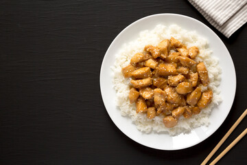 Homemade Orange Chicken with White Rice on a white plate on a black background, top view. Overhead, from above, flat lay. Space for text.