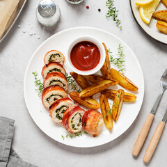 Bacon wrapped chicken rolls stuffed with feta cheese and spinach served with baked potato wedges and tomato sauce. Beautiful festive lunch or dinner. Gray stone background. top view	