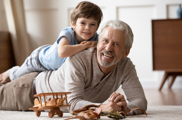 Portrait of happy mature Caucasian grandfather lying on floor at home play with little 8s grandson...