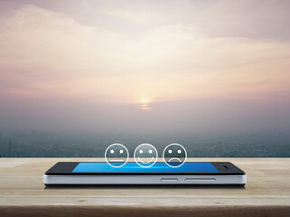 Excellent smiley face rating icon on modern smart mobile phone screen on wooden table over city tower and skyscraper at sunset, vintage style, Business customer service evaluation and feedback rating 
