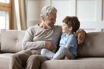 Loving old 60s Caucasian granddad sit on sofa with little curious grandson talk and chat at home. Caring mature grey-haired grandfather relax on couch with small grandchild gossip share secrets. - obrazy, fototapety, plakaty