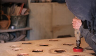 worker in dusty clothes with effort presses a drill with a crown bit on a sheet of thick plywood cutting round holes in the wood on a blurred background of the workshop, workflow in the carpentry room