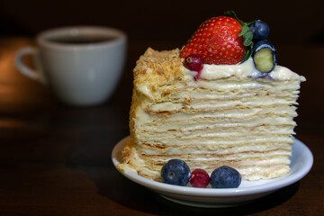 The piece of multi-layered cake decorated with mixed berries. Blueberries, strawberries, redcurrant on small plate and cup of tea or coffee on brown background. Soft selective focus, low key photo.