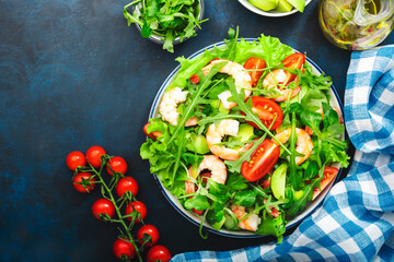 Fresh shrimp salad with tomatoes, lettuce, arugula, avocado, cucumber and lemon dressing on blue background. Healthy eating, clean food concept. Top view