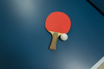 A red racket with a wooden handle for table tennis and a white ping-pong ball lie on a shady blue table top view