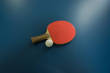 A red racket with a wooden handle for table tennis and a white ping-pong ball lie on a shady blue table top view