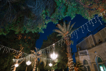 Crystal chandeliers hang from a garland between palm trees at night. Decorating an open-air wedding dinner.