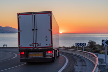 Truck with refrigerated semi-trailer on a curve of a mountain road by the sea and with the sunrise sun in the background.